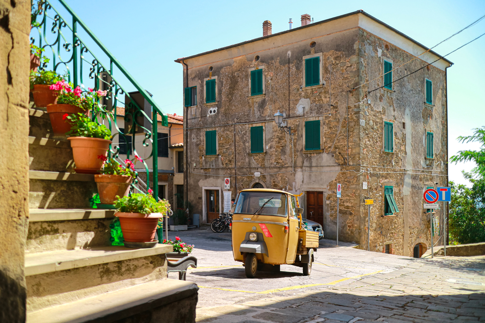 Castiglione della Pescaia, Vetulonia, Italy - 07/23/2020: Three wheeled car Ape Piaggio parked in the small square of an old italian village in Tuscany, with small plants on the stairs and old houses