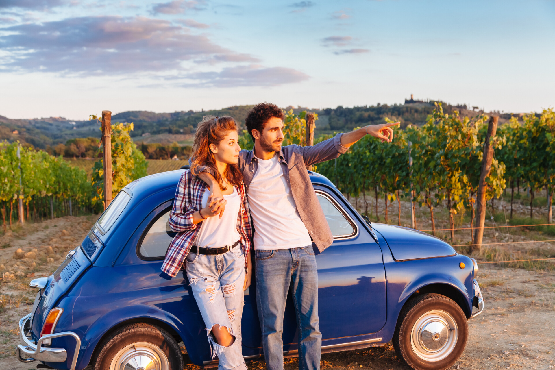 Kochająca para oglądać zachód słońca, stojąc opierając się o stary niebieski samochód w Toskanii we Włoszech. Wokół nich rzędy winorośli i klasyczne wzgórza. Młody człowiek pokazuje jego dziewczyna zachód słońca