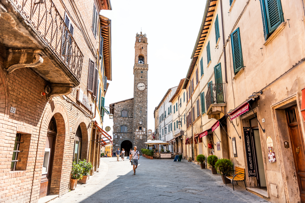 Montalcino, Italy - August 26, 2018: Street in town village in Tuscany during summer day and clock bell tower with people walking and stores in alley